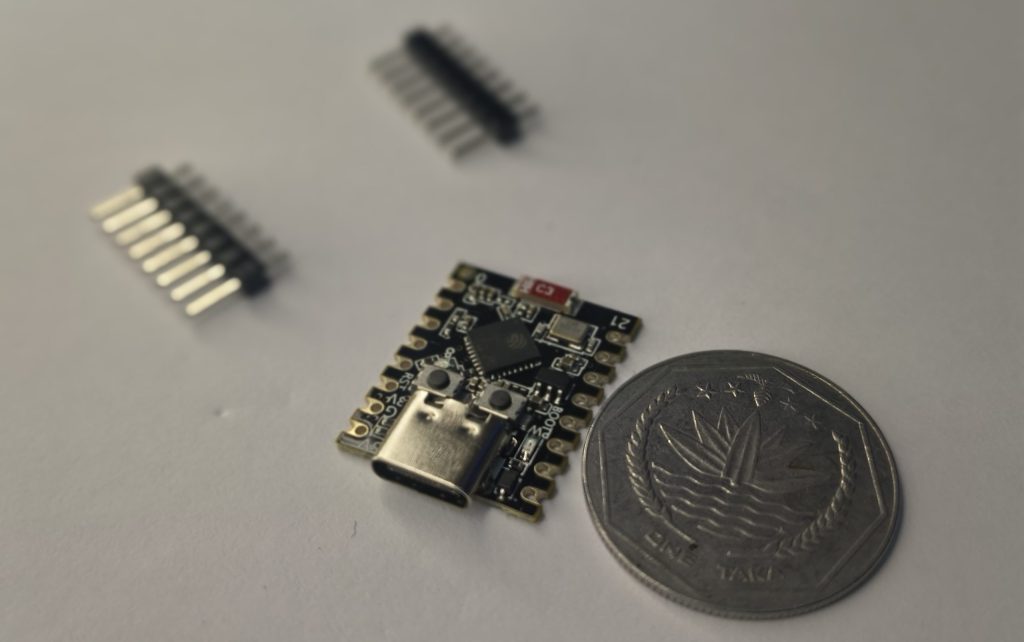 A close-up, high-angle photo of a black ESP32-C3 SuperMini microcontroller board with a USB-C port, sitting next to a silver Bangladeshi One Taka coin for scale. Two sets of header pins lie blurred in the background on a white surface.