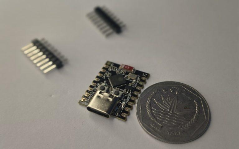 A close-up, high-angle photo of a black ESP32-C3 SuperMini microcontroller board with a USB-C port, sitting next to a silver Bangladeshi One Taka coin for scale. Two sets of header pins lie blurred in the background on a white surface.