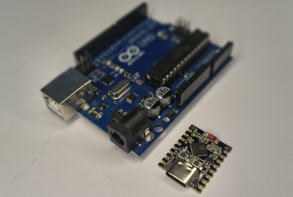 A top-down, slightly angled photo of a blue Arduino Uno R3 placed next to a much smaller, black Seeed Studio XIAO ESP32C3 microcontroller on a white surface.