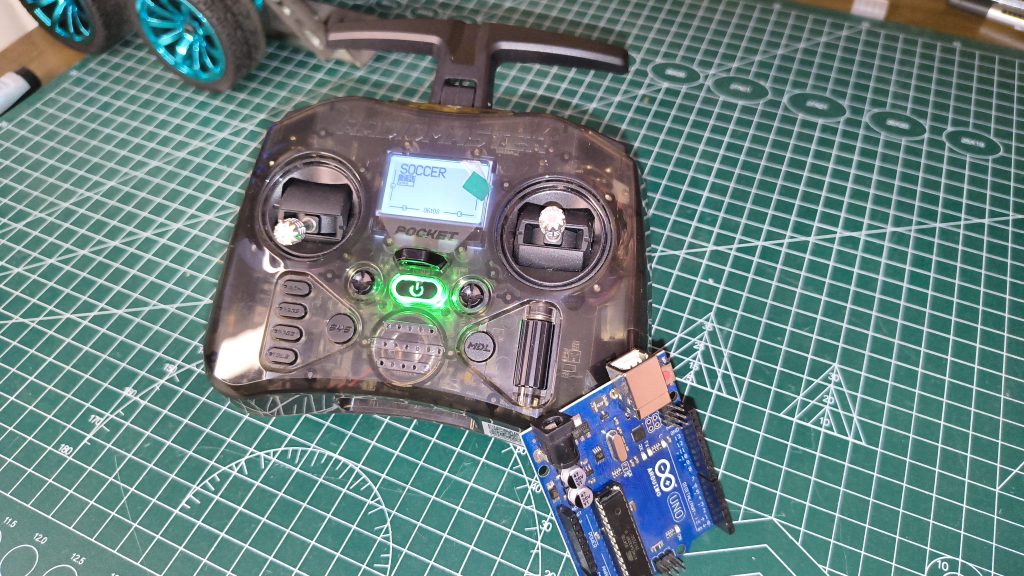 Top-down view of a RadioMaster Pocket radio showing the "SOCCER" model profile on screen, positioned alongside an Arduino Uno board on a workbench with robot wheels in the background.