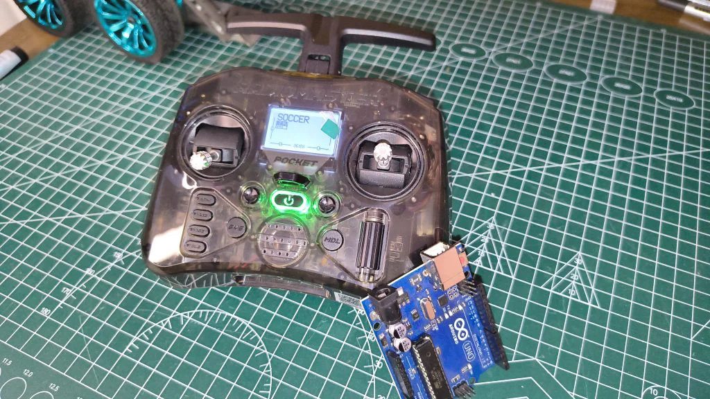 Top-down view of a RadioMaster Pocket radio showing the "SOCCER" model profile on screen, positioned alongside an Arduino Uno board on a workbench with robot wheels in the background.