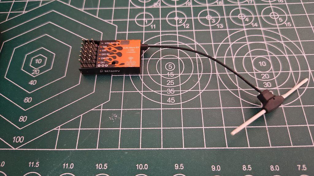 A BETAFPV ELRS Micro RX 5-channel 2.4GHz receiver with an orange and black flame design, connected to a black and white T-style dipole antenna on a green self-healing cutting mat.