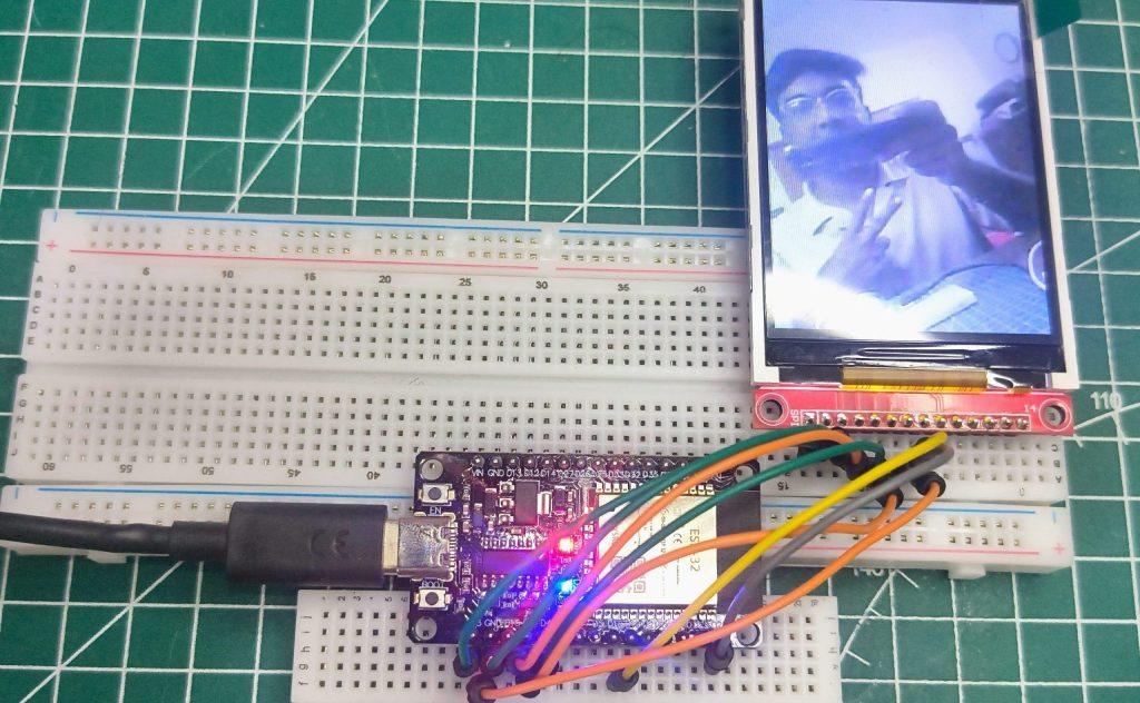 Top-down view of an ESP32 development board wired to a 2.8-inch TFT LCD display on a white breadboard. The screen displays a live video feed of a person, and the setup is placed on a green self-healing cutting mat with grid lines.