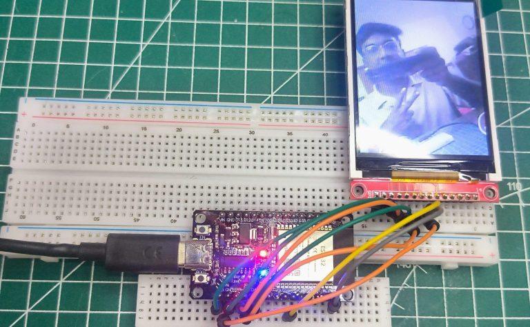 Top-down view of an ESP32 development board wired to a 2.8-inch TFT LCD display on a white breadboard. The screen displays a live video feed of a person, and the setup is placed on a green self-healing cutting mat with grid lines.