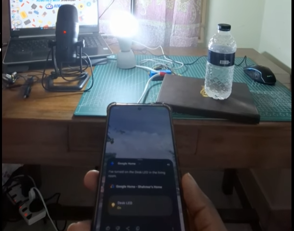 A person holding a smartphone displaying the Google Home app interface, which shows a "Desk LED" is turned on. In the background, a bright LED bulb is illuminated on a wooden desk next to a laptop, a professional microphone, and a green cutting mat.