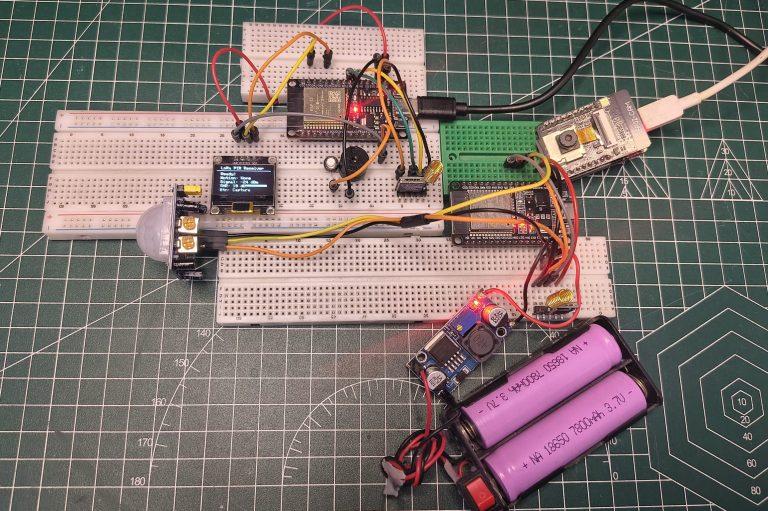Title Prototyping a LoRa-Based Wireless Security and Surveillance System Alt Text A high-angle, top-down photograph of an electronics project assembled on three white breadboards over a green cutting mat. The circuit features two ESP32 microcontrollers, one of which is an ESP32-CAM module, and two LoRa modules with gold coil antennas. A PIR motion sensor is connected to the left side, and a small OLED display shows technical data such as "Motion: None" and "Signal: -24 dBm." The system is powered by a battery holder containing two purple 18650 Li-ion batteries connected to a blue buck converter module. Jumper wires in various colors (orange, yellow, red, black) connect the components.