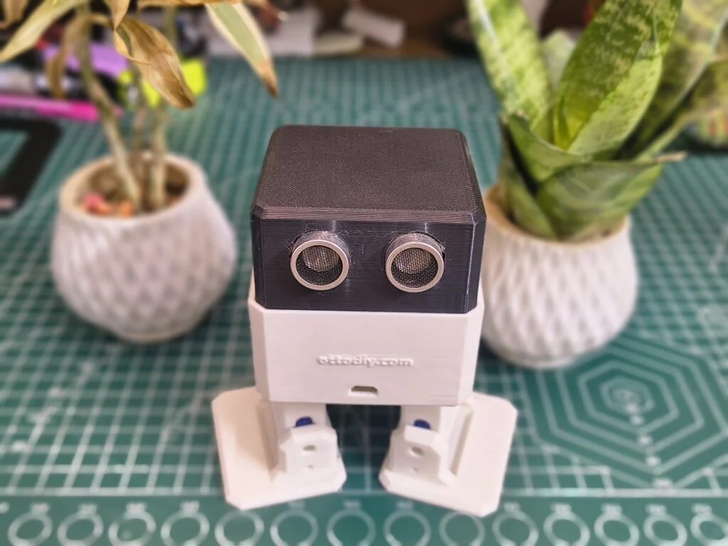 A small, 3D-printed Otto DIY bipedal robot with a black head and white body standing on a green cutting mat. The robot features two large ultrasonic sensor "eyes" for obstacle avoidance. It is positioned between two small potted plants, highlighting its compact size and friendly design.