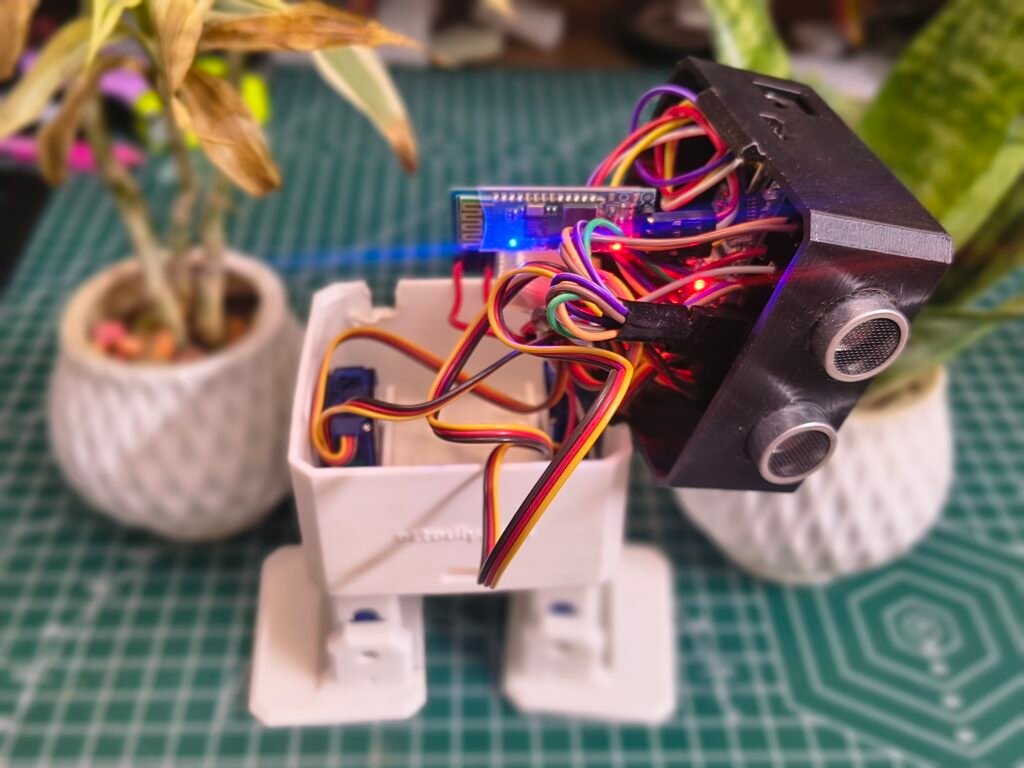 An Otto DIY robot with its black head opened to reveal a dense network of multi-colored jumper wires connected to an internal microcontroller. A Bluetooth module with a glowing blue LED is visible among the electronics. The robot's white bipedal base and ultrasonic sensor "eyes" are also shown. The setup is resting on a green cutting mat between two small potted plants.