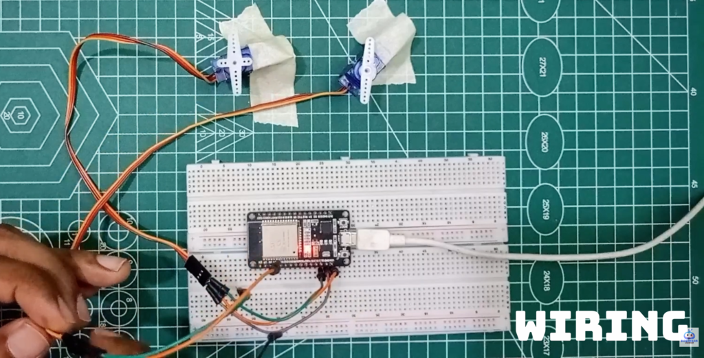 A top-down view of a green cutting mat featuring an ESP32 development board on a white breadboard. Two blue SG90 micro servo motors are secured with tape at the top, with their wiring connected to the ESP32. A white USB cable powers the board, and a hand is visible at the bottom left adjusting a connection. The text "WIRING" is overlaid in the bottom right corner.