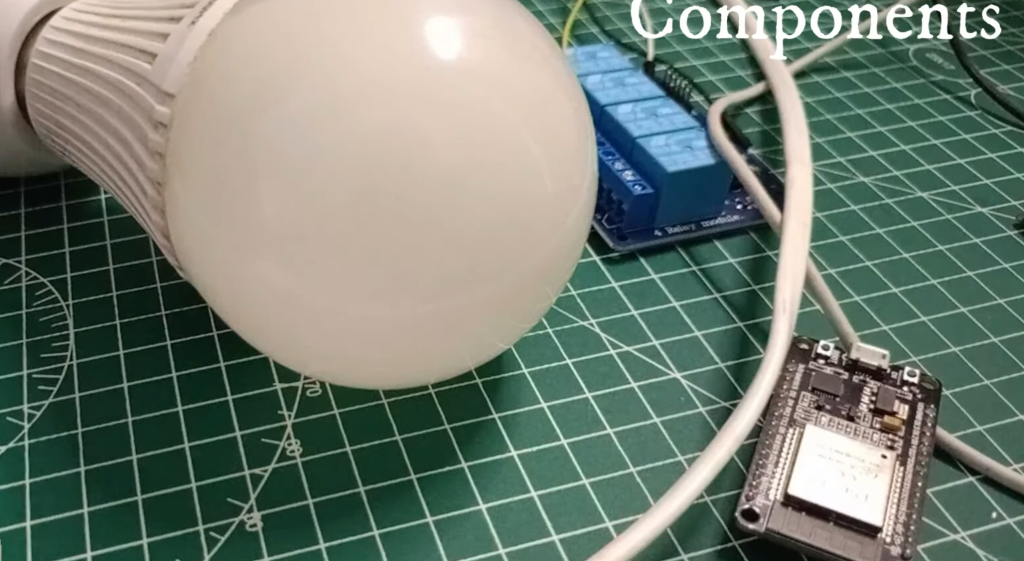 Close-up of components for a DIY electronics project on a green cutting mat, including a white LED light bulb on a socket base, a blue 4-channel relay module, and a black ESP32 microcontroller development board. The word "Components" is visible in the top-right.