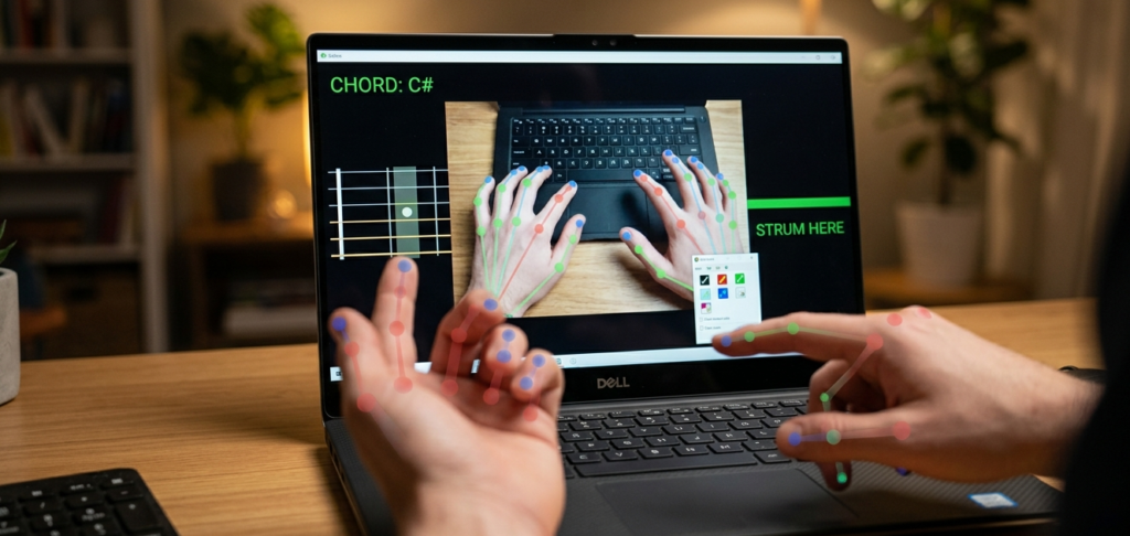 A first-person view of a developer using a Dell laptop to run an "Air Guitar" program. On the screen, a real-time camera feed tracks the user's hands with colored skeletal overlays. The software interface displays "CHORD: C#" and a virtual fretboard on the left, with a "STRUM HERE" indicator on the right. The user's physical hands are in the foreground, mirroring the digital tracking.