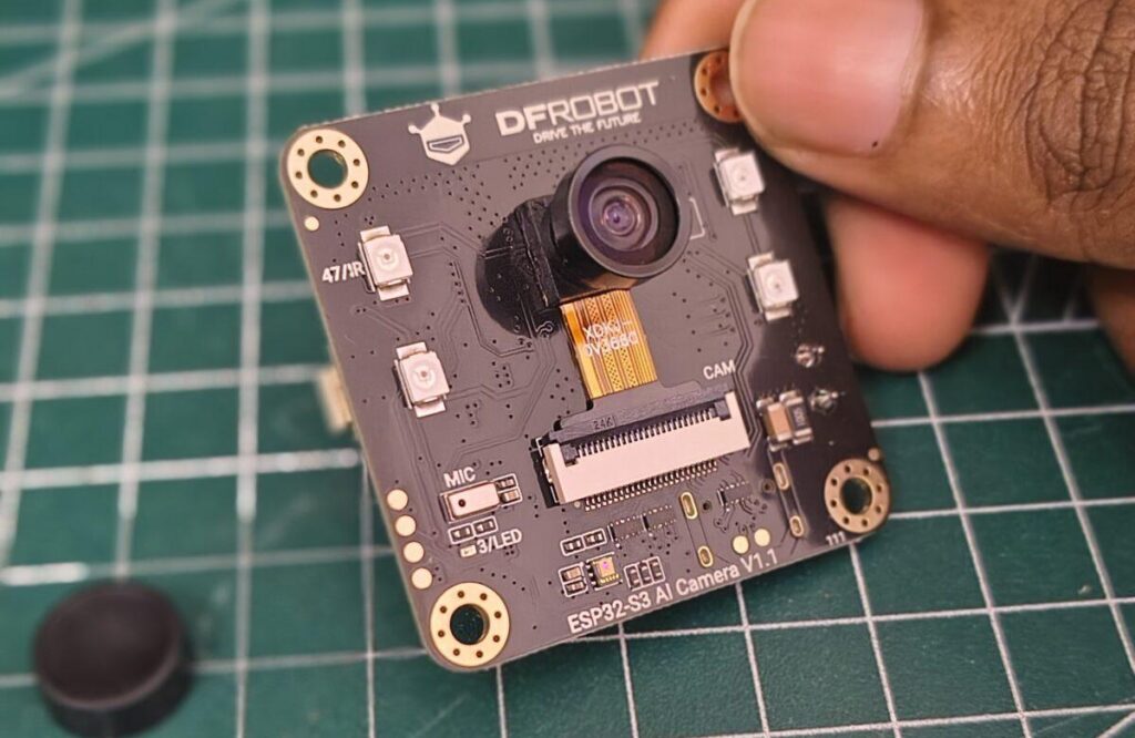 A close-up view of a person holding the DFRobot ESP32 S3 AI Camera V1.1 development board over a green cutting mat. The black PCB features a central camera sensor labeled OV3660 attached via a ribbon cable, surrounding IR/LED lights, a built-in microphone, and mounting holes.