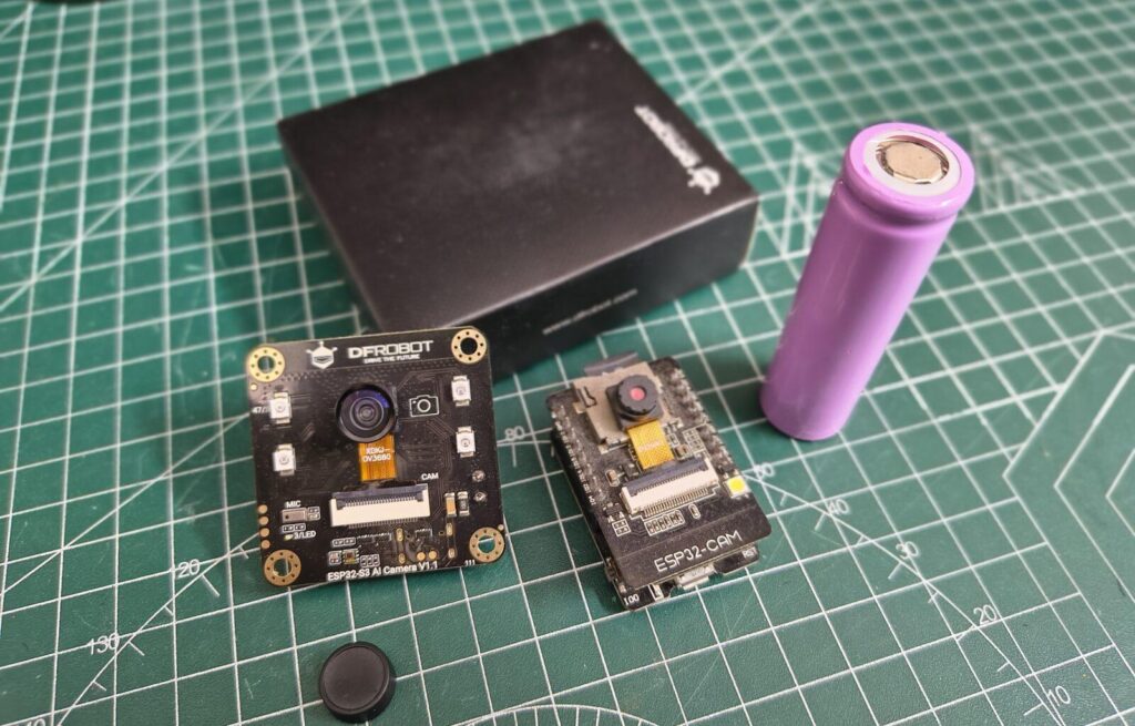 A top-down comparison shot on a green grid cutting mat featuring the DFRobot ESP32 S3 AI Camera V1.1 on the left and a standard ESP32-CAM module on the right. A purple 18650 lithium-ion battery stands vertically beside them next to a black DFRobot product box, showcasing the relative size differences of the hardware components.