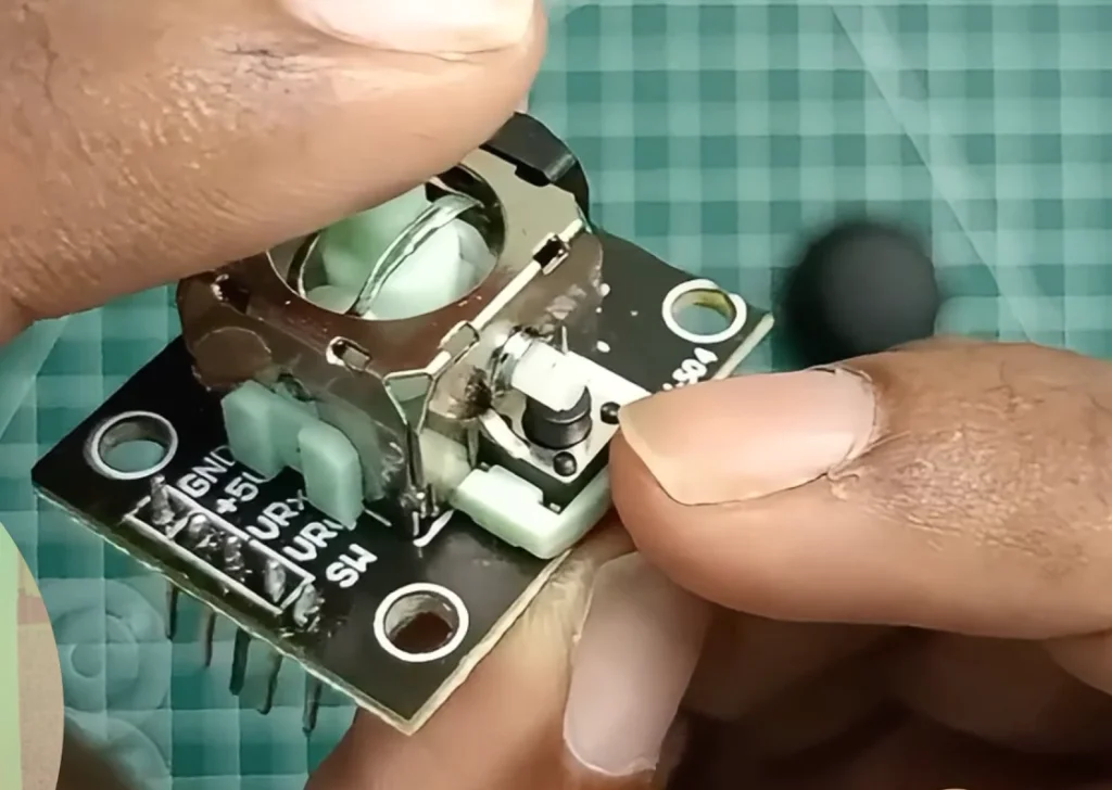 An extreme close-up view of two hands carefully holding a disassembled analog joystick module over a green grid mat. The black thumbstick cap is off to the side, exposing the white mechanical center and the two green side potentiometers. Clear pin labeling for GND, +5V, VRx, VRy, and SW is visible on the PCB edge.