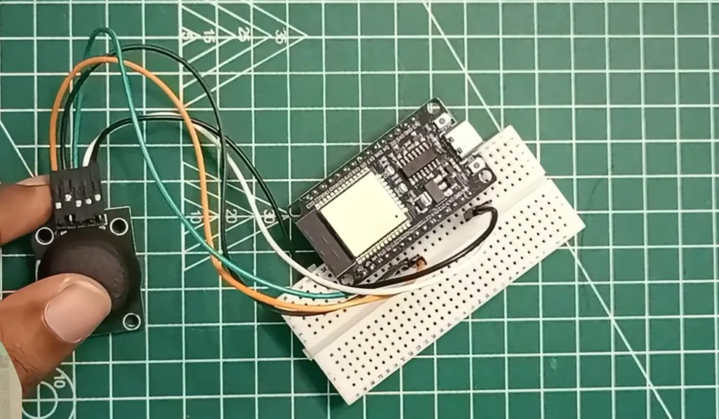 A top-down view on a green cutting mat showing a black analog joystick module being held by a thumb, connected via four jumper wires (orange, green, black, white) to an ESP32 development board. The ESP32 is mounted on a small white breadboard, illustrating a clean prototype setup for a Joystick with ESP32 project.