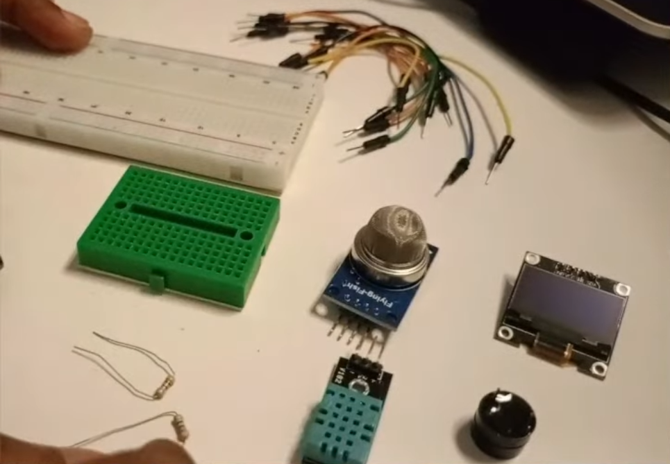 A collection of electronics components for a diy weather station project laid out on a white surface. The set includes a large white breadboard, a small green breadboard, an MQ gas sensor, a DHT temperature and humidity sensor, an OLED display module, a small buzzer, resistors, and several multi-colored jumper wires.