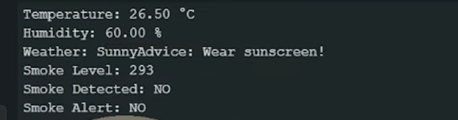 A digital readout from a diy weather station project showing environmental data. The text on a dark background displays a temperature of 26.50°C, humidity at 60.00%, weather as "Sunny," and an advice note saying "Wear sunscreen!". Below this, it lists a smoke level of 293 with "Smoke Detected: NO" and "Smoke Alert: NO".