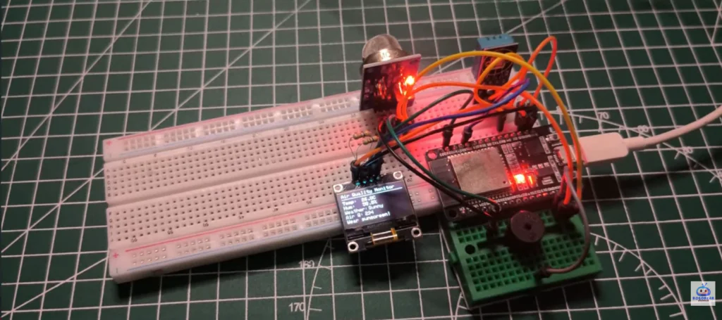 Build Your Own ESP32 DIY Weather Station: Track Temperature, Humidity & Air Quality 3 A top-down view of a DIY weather station prototype built on a breadboard over a green grid mat. The setup features an ESP32 microcontroller, an MQ gas sensor with a glowing red LED, a blue temperature sensor, and a small OLED display showing "Air Quality Monitor" with live temperature and humidity data. A white USB cable provides power to the system.